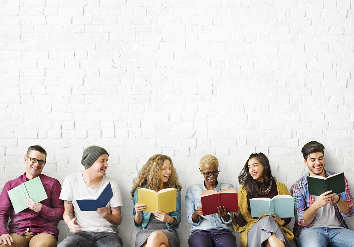line of people with books sitting against a brick wall