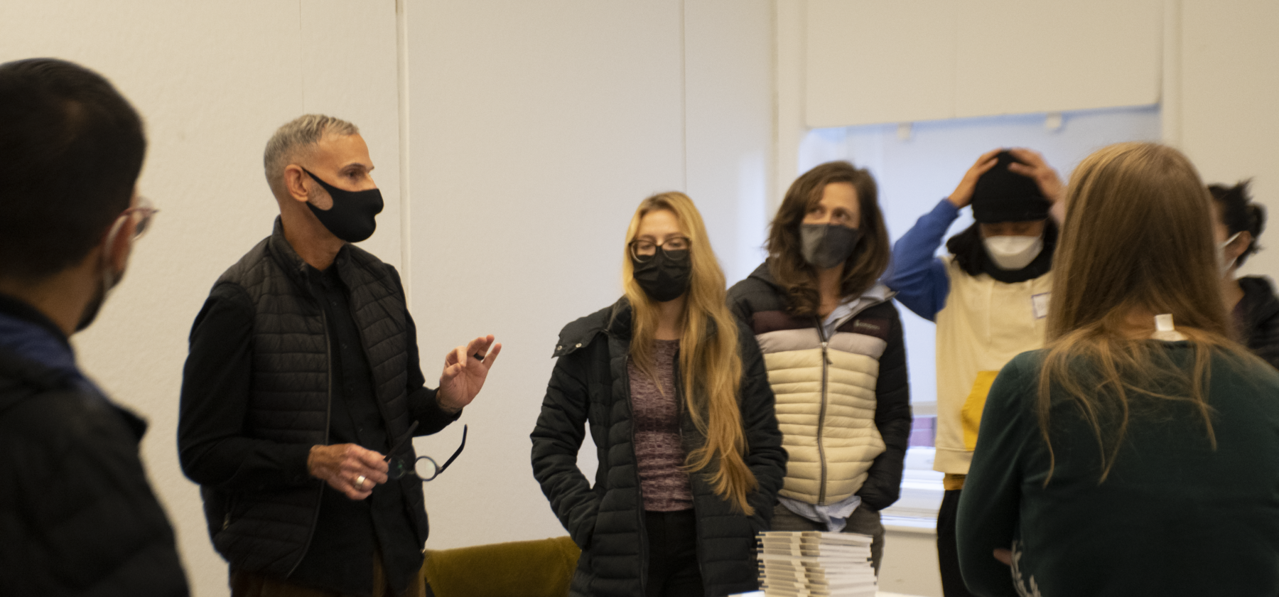 Ernesto Pujol is standing in the middle of a room, surrounded by 6 people looking at him as he is speaking. Everyone in the room has a mask on and Pujol has his hands raised at his waist.