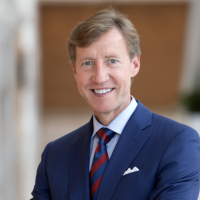 Headshot of man with light brown hair and light skin smiling looking at the camera. Wearing a blue suit with a red and blue strip tie.