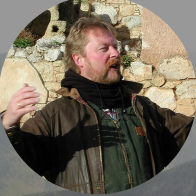 Profile view photo of male figure with light skin and short reddish hair speaking with arms raised wearing green top and brown leather coat, with black scarf