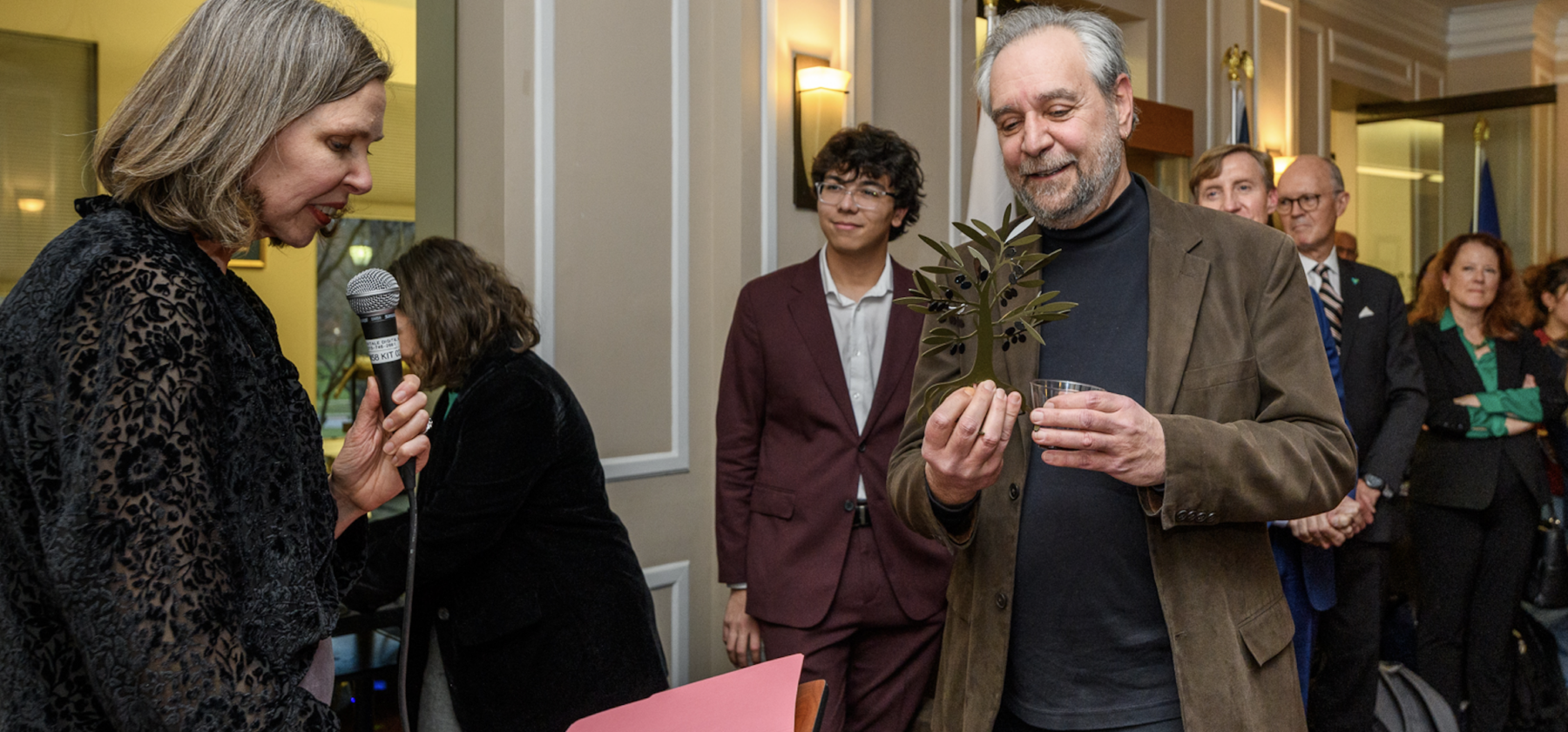 Award presented to honoree by speaker holding microphone. Recipient accepting olive tree sculpture.