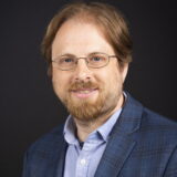 headshot of man with red hair, beard and mustache wearing a light blue button down shirt and blue checkered blazer