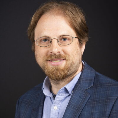 headshot of man with red hair, beard and mustache wearing a light blue button down shirt and blue checkered blazer