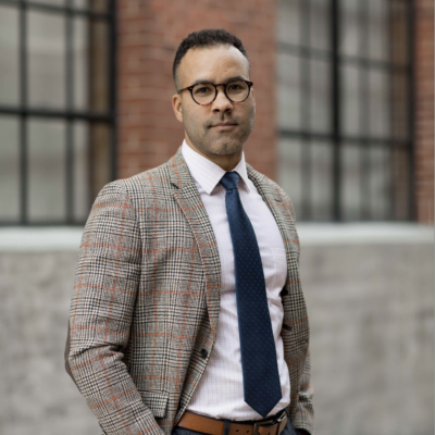 male figure with short dark hair wearing glasses and facing camera with slight smile. Wearing blazer jacket and tie