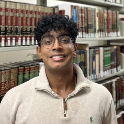 Man with dark short hair smiling at camera standing in front of book shelves wearing beige sweater