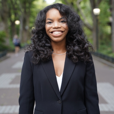 Woman with long black standing outdoors smiling at the camera wearing a black blazer and white shirt