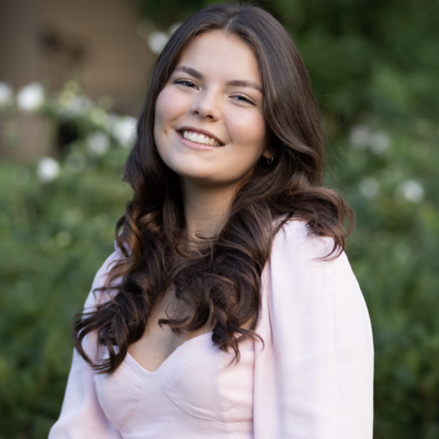 Woman with long brown hair smiling at camera outdoors wearing light pink dress in front of green tree with white flowers