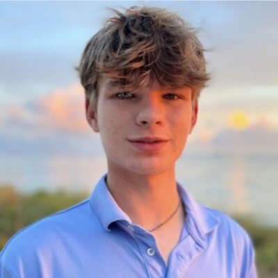 Headshot of man with blonde short hair smiling at the camera standing outdoors with water, clouds, and greenery in the background. He is wearing a light blue button down shirt.