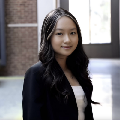 Headshot of woman with long dark hair smiling at the camera in front of brick wall with window wearing a black blazer and white shirt