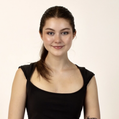 Headshot of woman with long brown hair pulled back in a pony tail smiling at the camera against an off-white solid background wearing a black short sleeve top