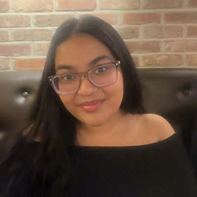 Headshot of woman with long black hair and glasses smiling at the camera against a brick background a black leather chair