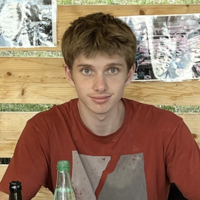 Mam with short brown hair smiling at camera seated against a wooden slat background and wearing a rust colored short sleeve shirt