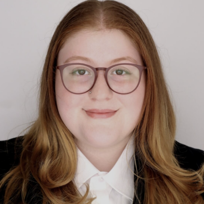 Woman with long red hair and glasses smiling at the camera against an off white background wearing a black blazer and white shirt