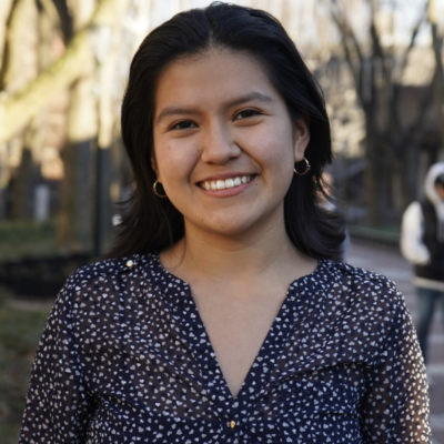 Heashot of woman with long black hair smiling standing outdoors on campus wearing a black and white dotted blouse