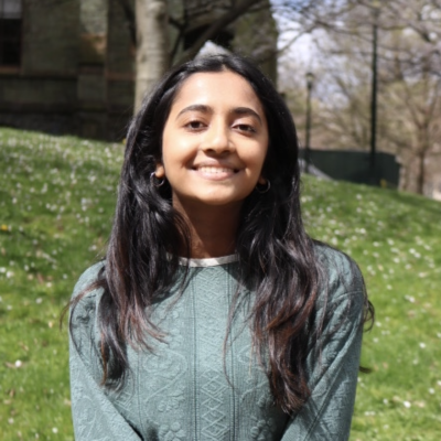 Headshot of woman with long black hair standing outdoors smiling at camera against a grassy background wearing a light green sweater