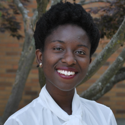 Female masters student with short black hair and dark skin smiling at camera wearing white blouse standing outdoors with a tree in background
