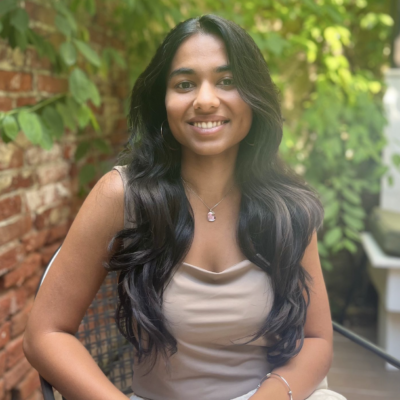 Female doctoral student with long black hair and beige sleeveless top with brown skin sitting outside with greenery in background smiling at camera