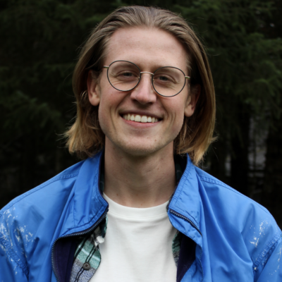 Male doctoral student with shoulder-length dirty blonde hair, light skin, and round glasses wearing a light blue jacket and white shirt standing outdoors