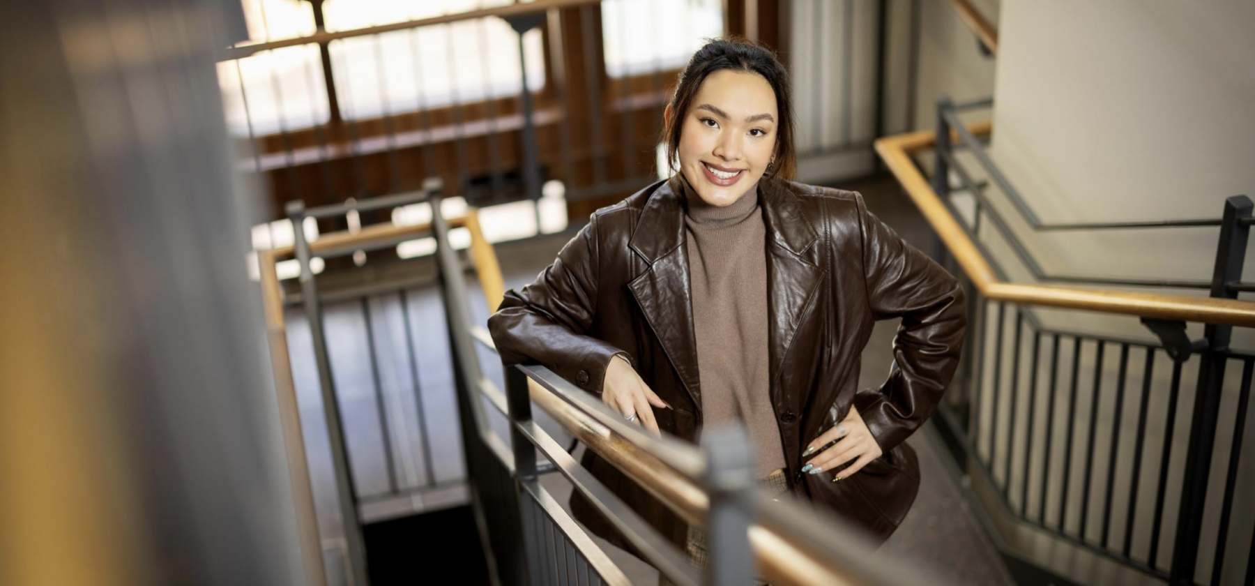 photo of program fellow on stairs smiling up at camera. she has light skin and brown hair pulled back and is wearing a brown leather jacket