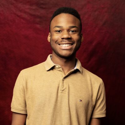 headshot of SNF Paideia Fellow Isaiah Littlejohn. College student with dark skin wearing a tan short sleeve shirt standing again a maroon background