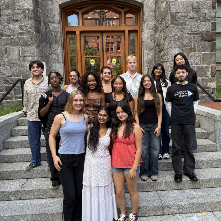 15 students gathered outside for a group photo on steps in front of stone campus building